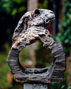 A rustic sculpture of a rabbit's head emerging from a circular base, set in a green outdoor environment.