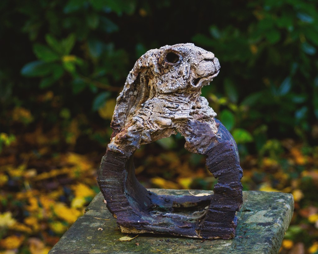 A textured sculpture of a rabbit's head, mounted on a circular base, set against a blurred background of green foliage and fallen leaves.