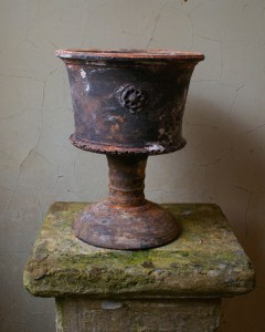 An aged stone pedestal supporting a weathered, ornate metal pot with a decorative handle.