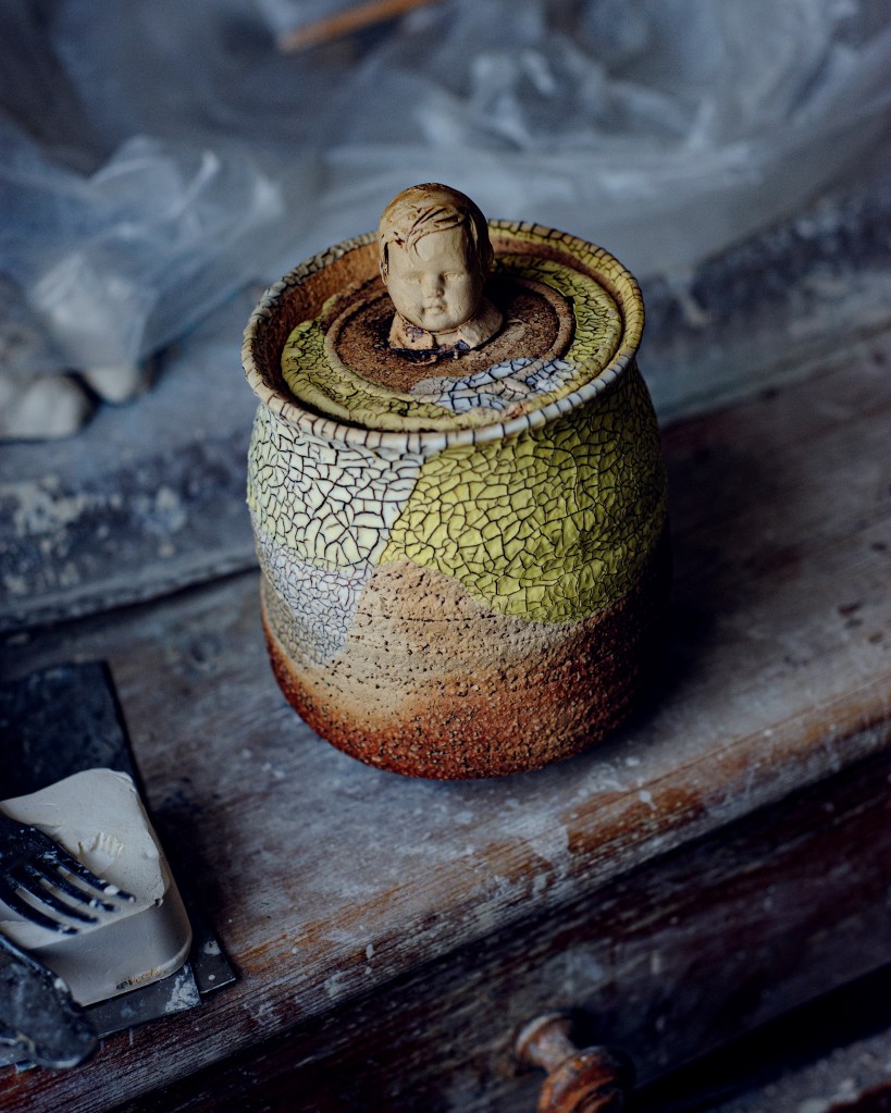 Eine keramische Dose mit einem Kopf aus Ton auf dem Deckel, auf einer Holzoberfläche, umgeben von Töpferwerkzeugen.