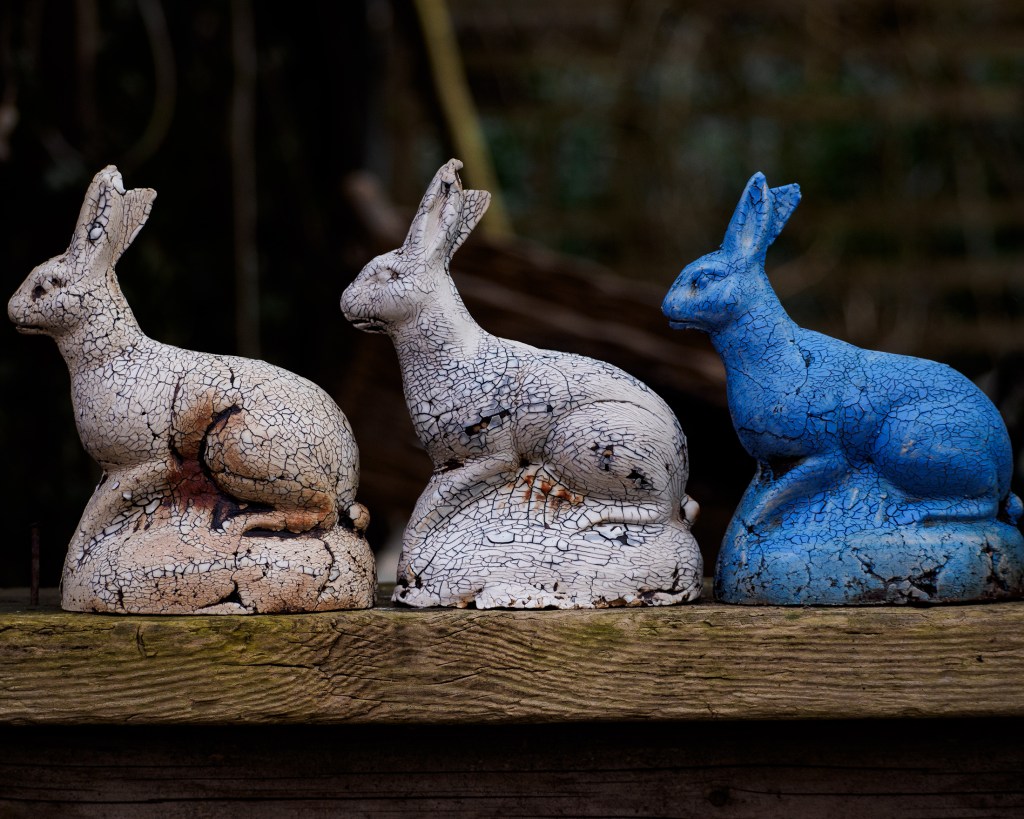 Drei dekorative Kaninchenstatuen in verschiedenen Farben: creme, weiß mit Rissen und blau, stehen auf einer Holzoberfläche.